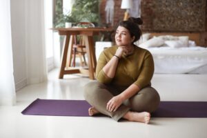 plus-size-woman-sitting-on-yoga-mat-on-floor-of-bedroom